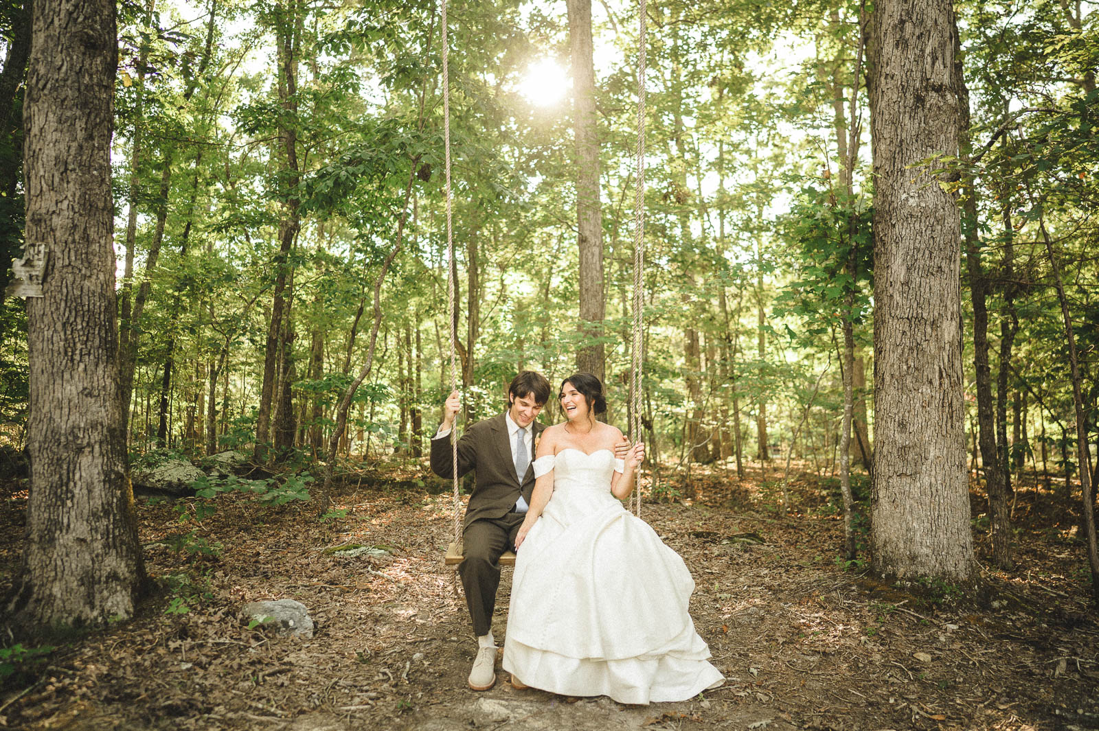 A bride and groom are on a wooden rope, swing in the middle of the woods with the sun shining behind them